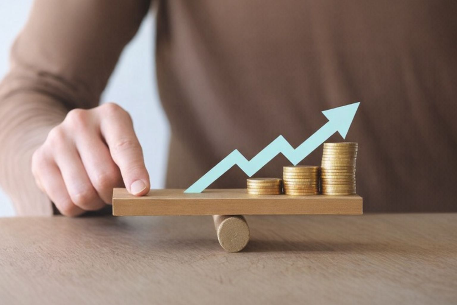 En hand balanserar en träplanka med myntstaplar och en stigande blå pil.